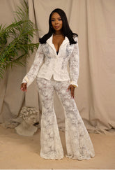 Woman wearing a white lace outfit standing against a beige curtain with a plant and sculpture in the background.