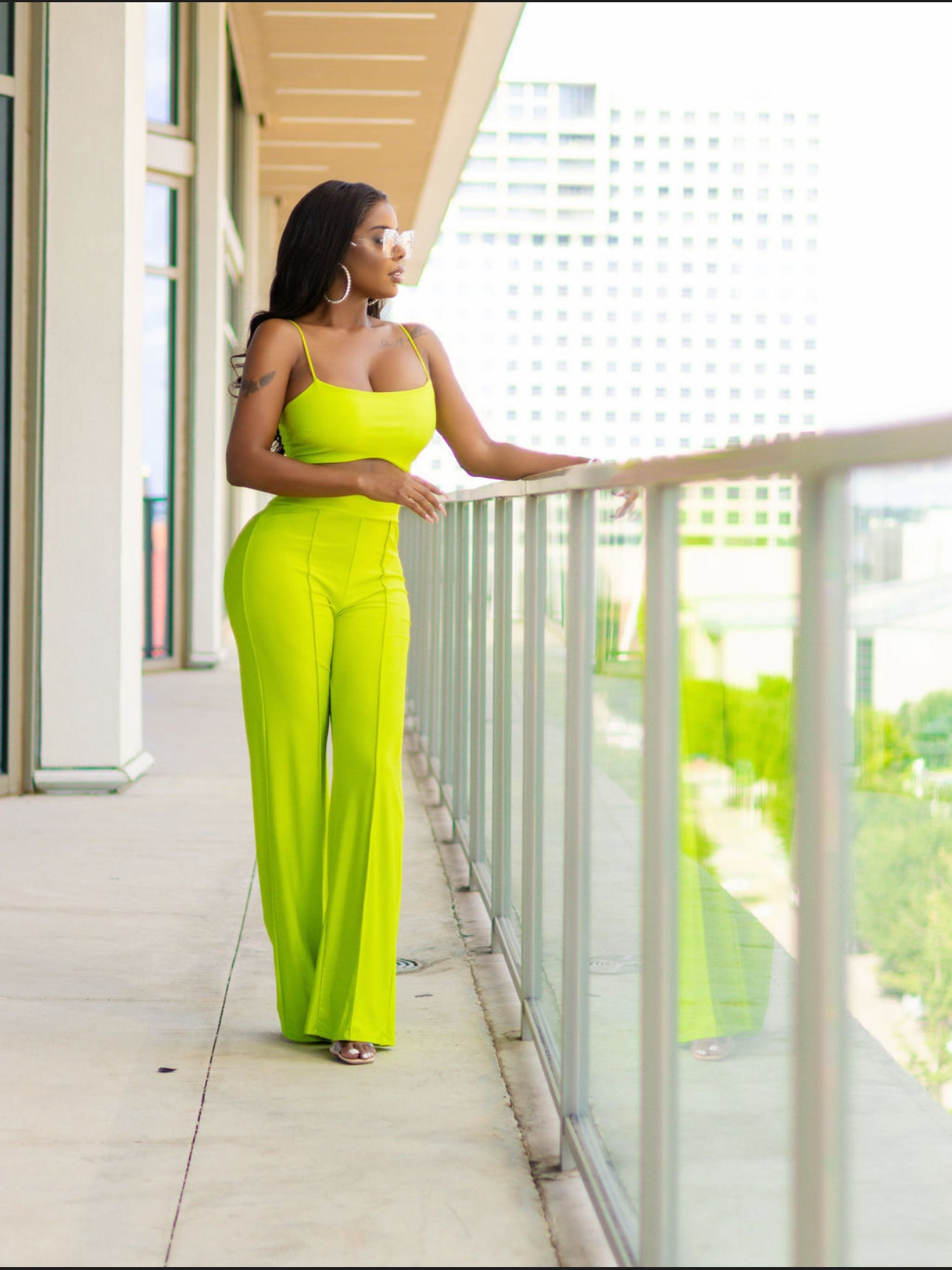 Woman in a bright green jumpsuit standing on a balcony with a cityscape view.