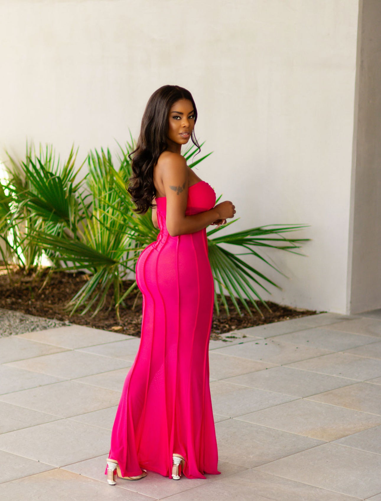 Woman in a bright pink dress standing outdoors with plants and a white wall in the background
